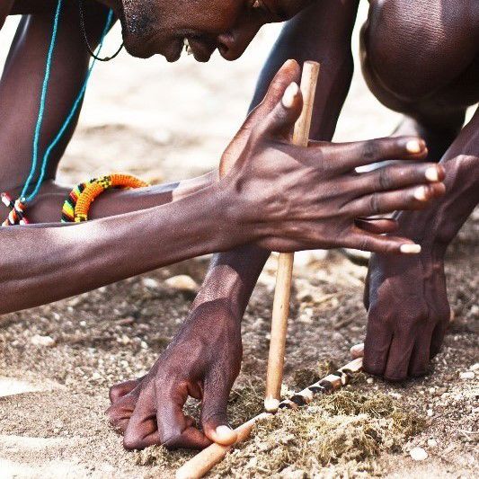 A photograph of people in traditional African dress lighting a fire using two wooden sticks on the ground.