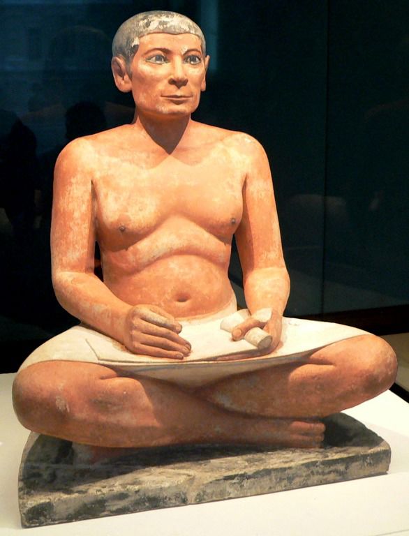 An Egyptian sculpture showing a scribe sitting cross-legged on the ground with a roll of papyrus.
