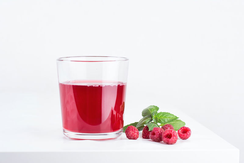 Photograph of rasperries and rasperry juice in a glass on a white background.