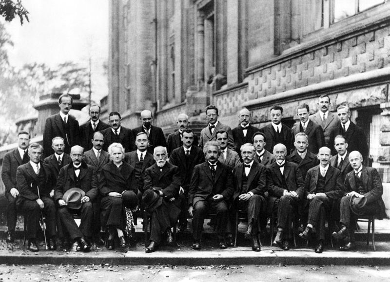 Black and white photograph of people in suits standing and sitting outside a large building. Top row, left to right: A. Piccard, E. Henriot, P. Ehrenfest, E. Herzen, Th. de Donder, E. Schrödinger, J.E. Verschaffelt, W. Pauli, W. Heisenberg, R.H. Fowler, L. Brillouin. Middle row, left to right: P. Debye, M. Knudsen, W.L. Bragg, H.A. Kramers, P.A.M. Dirac, A.H. Compton, L. de Broglie, M. Born, N. Bohr. Bottom row, left to right: I. Langmuir, M. Planck, M. Curie, H.A. Lorentz, A. Einstein, P. Langevin, Ch.-E. Guye, C.T.R. Wilson, O.W. Richardson